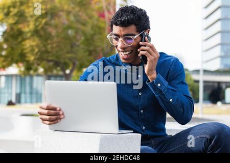Adult ethnic cheerful male remote employee having a conversation on smartphone while working on netbook in town on sunny day Stock Photo