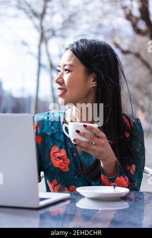 Long-haired brunette Asian Woman having a coffee on a terrace of a cafe while is working with a laptop Stock Photo