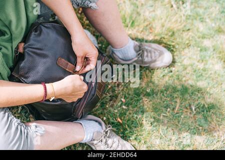 High angle crop anonymous female in casual wear sitting on grass and closing leather backpack while camping in sunny nature Stock Photo