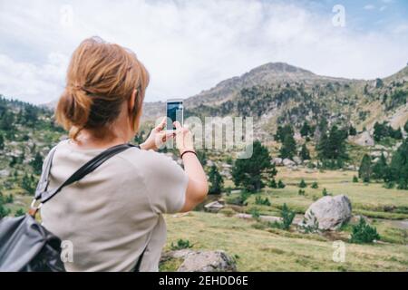 Back view anonymous female backpacker taking pictures on smartphone of amazing stony verdant highlands in Ruda Valley in Catalan Pyrenees Stock Photo