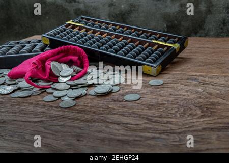 Vintage wooden hand abacus with pile of money coins from red cloth bag on rustic old wooden table. Concept for making business and saving money of chi Stock Photo
