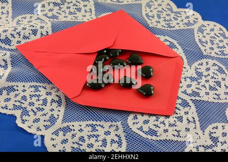 Black hearts spilling out of open red Valentine envelope on pink heart lace table runner on blue tablecloth Stock Photo