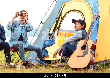 A group of friends camping near the lake. Women use binoculars to look at birds in the sky. Stock Photo
