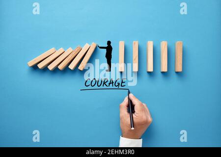 Businessman hand writing the word courage with silhouette of a man making a stop gesture to prevent wooden dominos from collapsing. Stock Photo