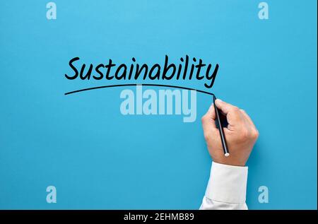 Businessman hand writing the word sustainability on blue background. Corporate environmental ecology strategy in business concept. Stock Photo