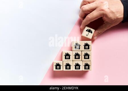 Male hand placing a protective umbrella icon on top of the wooden cubes with house icons. Real estate or house insurance concept. Stock Photo