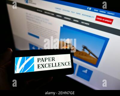 Person holding mobile phone with logo of Canadian pulp manufacturer Paper Excellence B.V. on screen in front of web page. Focus on cellphone display. Stock Photo