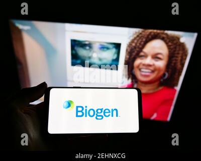 Person holding mobile phone with business logo of American biotechnology company Biogen Inc. on screen in front of web page. Focus on phone display. Stock Photo