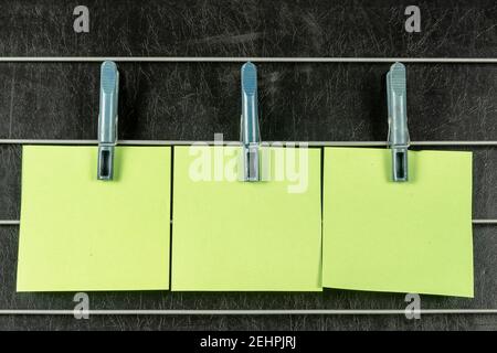The paper sticky notes hanging from clothespins. Three blank light green square stickers on a black background. Stock Photo