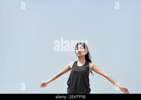 young asian woman in sportswear standing outdoors against blue sky with open arms Stock Photo