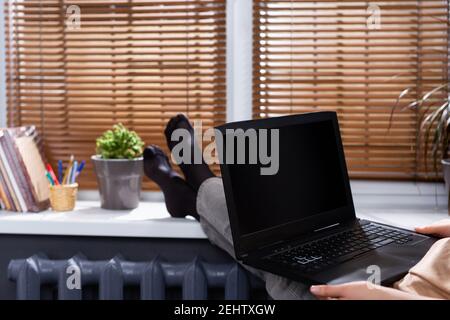 Cropped photo of a female student with her feet on the windowsill at home, holding a laptop with a black screen. Online training. Stock Photo
