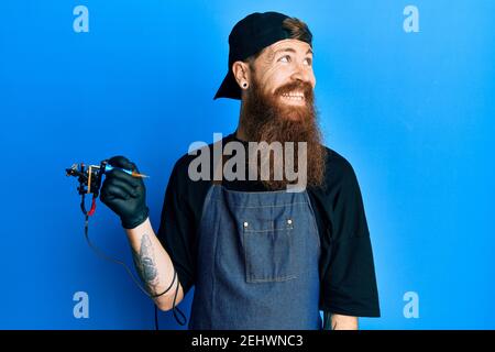 Redhead man with long beard tattoo artist wearing professional uniform and gloves smiling looking to the side and staring away thinking. Stock Photo