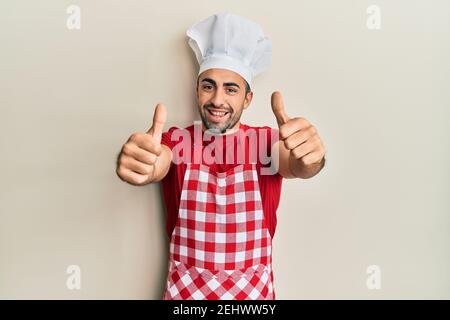 Young hispanic man wearing baker uniform approving doing positive gesture with hand, thumbs up smiling and happy for success. winner gesture. Stock Photo