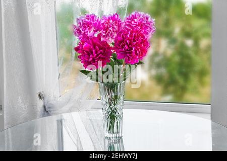 a bouquet of five pink flowers peonies standing in the room on the table in a crystal vase by the window Stock Photo