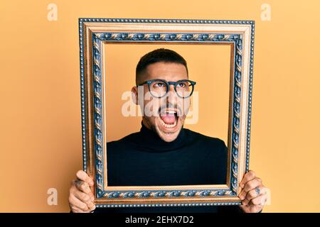 Handsome man with tattoos holding empty frame angry and mad screaming frustrated and furious, shouting with anger looking up. Stock Photo