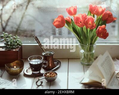 Oriental coffee cooked in traditional Turkish copper coffee pot with flowers on window sill. Wooden windowsill with bunch of tulips book.Cozy scene, h