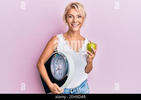 Young blonde girl holding weight machine and green apple smiling with a happy and cool smile on face. showing teeth. Stock Photo