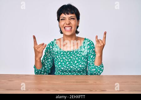 Beautiful brunettte woman wearing casual clothes sitting on the table shouting with crazy expression doing rock symbol with hands up. music star. heav Stock Photo