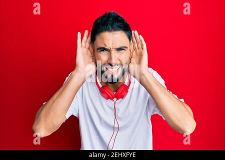 Young man with beard listening to music using headphones trying to hear both hands on ear gesture, curious for gossip. hearing problem, deaf Stock Photo