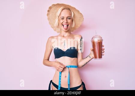 Young blonde woman with tattoo wearing bikini using tape measure and drinking smoothie sticking tongue out happy with funny expression. Stock Photo