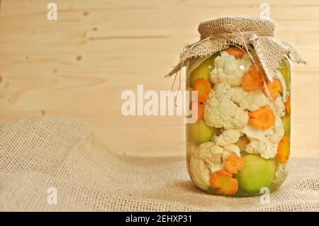 Concept of seasonal pickles jar covered with sackcloth full of colored green raw tomatoes, orange carrot and white cauliflower. Stock Photo
