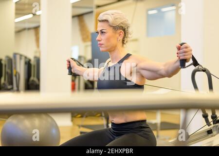 Sporty middle-aged woman working out on resistance equipment in a gym strengthening her arm and upper body core muscles viewed past other equipment in Stock Photo