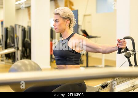 Sporty middle-aged woman working out on resistance equipment in a gym strengthening her arm and upper body core muscles viewed past other equipment in Stock Photo