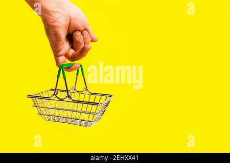 Hand holds shopping basket with its little finger, on yellow background. Concept of a trip to the supermarket. Stock Photo