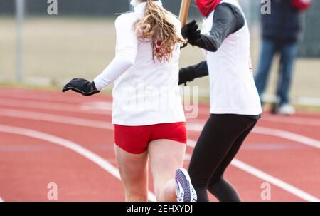 Girls exchanging the baton wearing gloves and long sleeves during a relay running race outdoors in the cold. Stock Photo