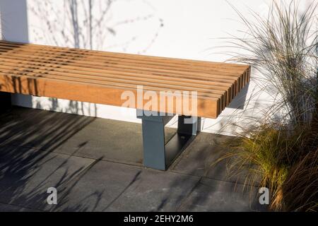 A modern wooden garden wood bench with metal frame legs seating area on a dark stone paved patio with white wall and ornamental grasses England UK GB Stock Photo