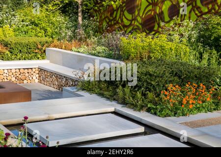 Modern English garden view Yorkstone stepping stone slabs over a water feature leading to a sunken garden with stone bench flower borders Spring UK Stock Photo