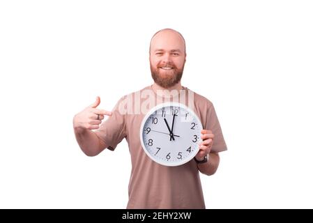 Cheerful smiling young bearded man pointing at white wall watch. Stock Photo