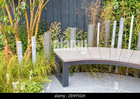 A modern garden bench on stone paved patio - planting of multi stemmed Prunus maackii Amber Beauty tree ornamental grasses - Deschampsia cespitosa UK Stock Photo