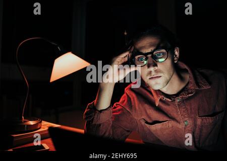 Caucasian male student working at laptop late at night tired leaning on hands meeting deadlines Stock Photo