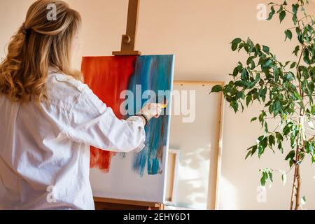 Woman working on contemporary abstract oil painting at creative studio. Blond hair caucasian painter standing with brush and easel indoors Stock Photo