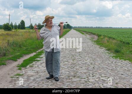 Outdoor portrait of Ukrainian senior farmer in straw hat standing on cobblestone road and taking walking stick Stock Photo