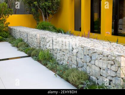 Mediterranean garden with stone filled gabion gabions wall, border of mixed herbs grey stone patio with a rill water feature England GB UK Stock Photo