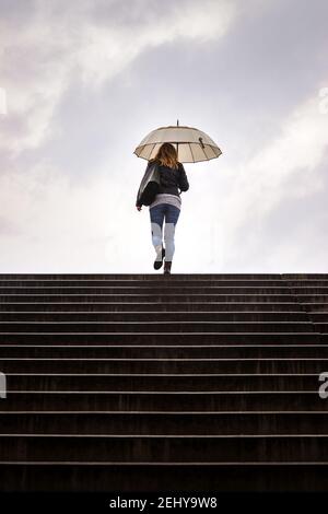 Stairway to heaven. Woman with umbrella walking at staircase in rain. Silhouette of woman against cloudy sky in city Stock Photo
