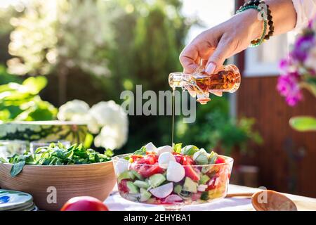 Woman hand pouring flavored olive oil to fresh vegetable salad. Preparation healthy vegetarian food for garden party outdoors Stock Photo