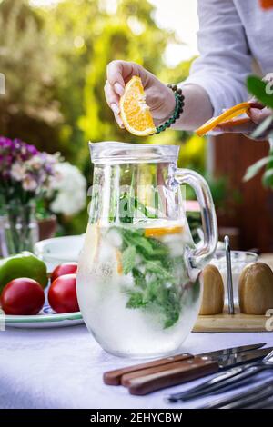 Sparkling water in carafe on table. Woman hand putting slice of orange into cold mineral water with mint and ice Stock Photo