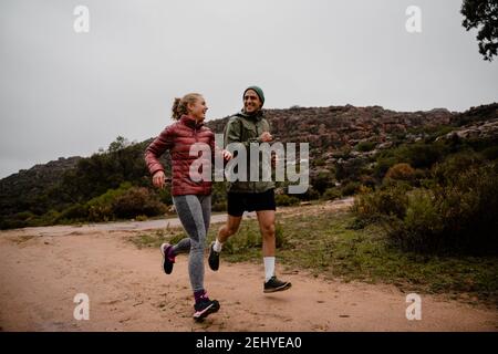 Young fit couple laughing while running on gravel mountain track in cloudy weather with luscious bushes Stock Photo