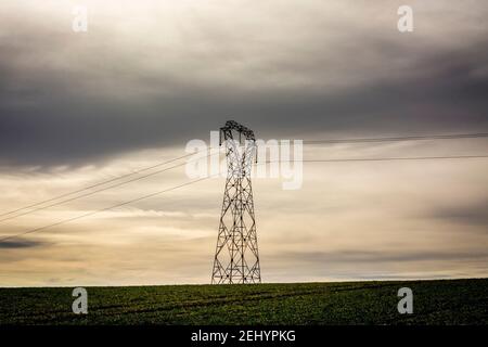 Power lines stand tall against a dramatic sky over a green field during the late afternoon Stock Photo