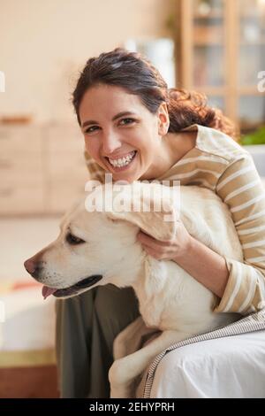 Vertical warm toned portrait of smiling young woman hugging dog sitting on bed in cozy home interior Stock Photo