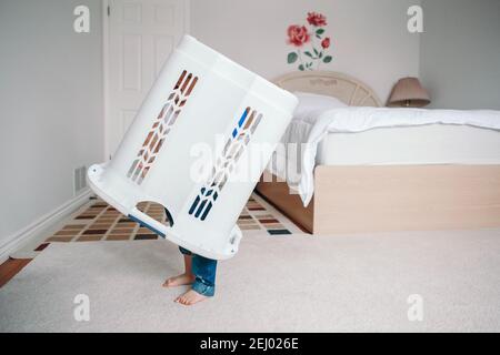 Cute funny baby toddler child with large clothes bin on head. Lonely autistic kid playing alone at home hide and seek game. Funny memorable childhood Stock Photo