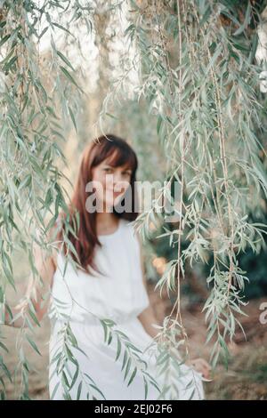 Blurry silhouette of Caucasian woman with long dark brown red hair among willow tree branches leaves outdoor. Casual style real woman female in long w Stock Photo