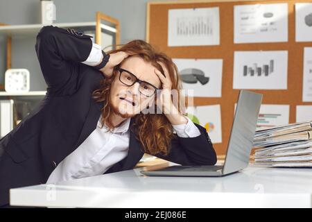 Funny young weird stressed man accountant sitting at laptop touching head feeling burnout Stock Photo
