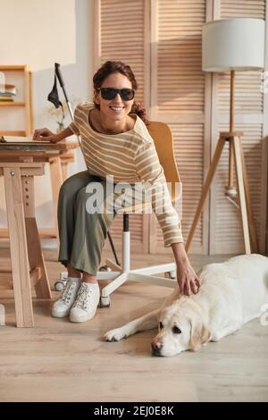Vertical full length portrait of modern blind woman sitting at table in cozy home interior and petting guide dog Stock Photo