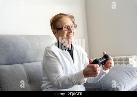 Glad senior female sitting on comfortable sofa in living room and playing video game while using gamepad Stock Photo