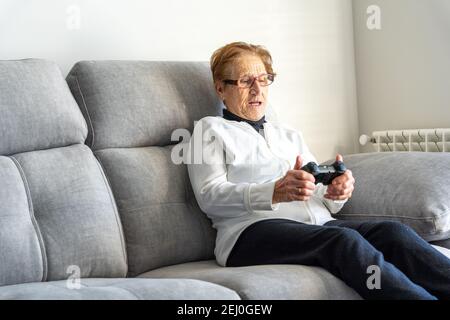 Delighted elderly female with console sitting on sofa and playing video game at home Stock Photo