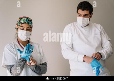 Male and female doctors in medical masks standing in hospital and putting on protective latex gloves while working during coronavirus pandemic Stock Photo